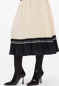 Pleated skirt in cream and black, with a contrasting black waistband featuring a logo. Black patent ankle boots with textured accents.