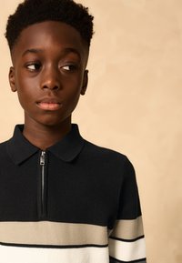 Polo negro con textura acanalada y cuello con cremallera que presenta rayas horizontales en beige y blanco. Textura suave con un diseño moderno y casual.