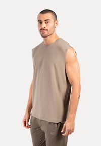 Muscle-fit ärmelloses T-Shirt aus beigem Stoff, runder Halsausschnitt, glatte Textur, mit genähten Kanten, getragen mit passenden Sporthosen.