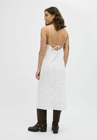 Vestido blanco con textura y patrones delicados, con tirantes finos y un escote bajo atado con una cinta. Combinado con botas marrones.