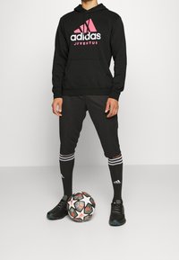 Felpa nera Adidas con logo rosa, pantaloni da jogging grigi, calzini neri con strisce bianche e scarpe da calcio nere accanto a un pallone da calcio a motivi.