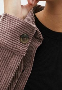 Chaqueta de pana con textura acanalada en rosa polvo, que presenta un cierre de botón redondo y una parte superior de color negro acanalada parcialmente visible por debajo.