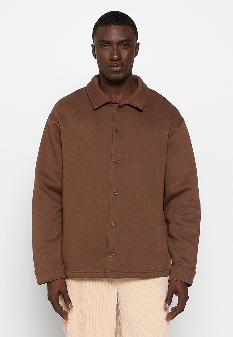 Veste marron à boutons avec un col évasé et des manches longues. Tissu doux et texturé. Coupe droite avec des détails de couture visibles.