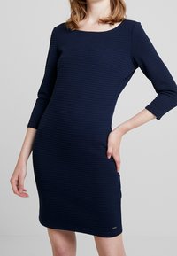 Robe bleu marine en tissu texturé, avec un col rond et des manches trois-quarts, silhouette ajustée et détail de marque subtil.