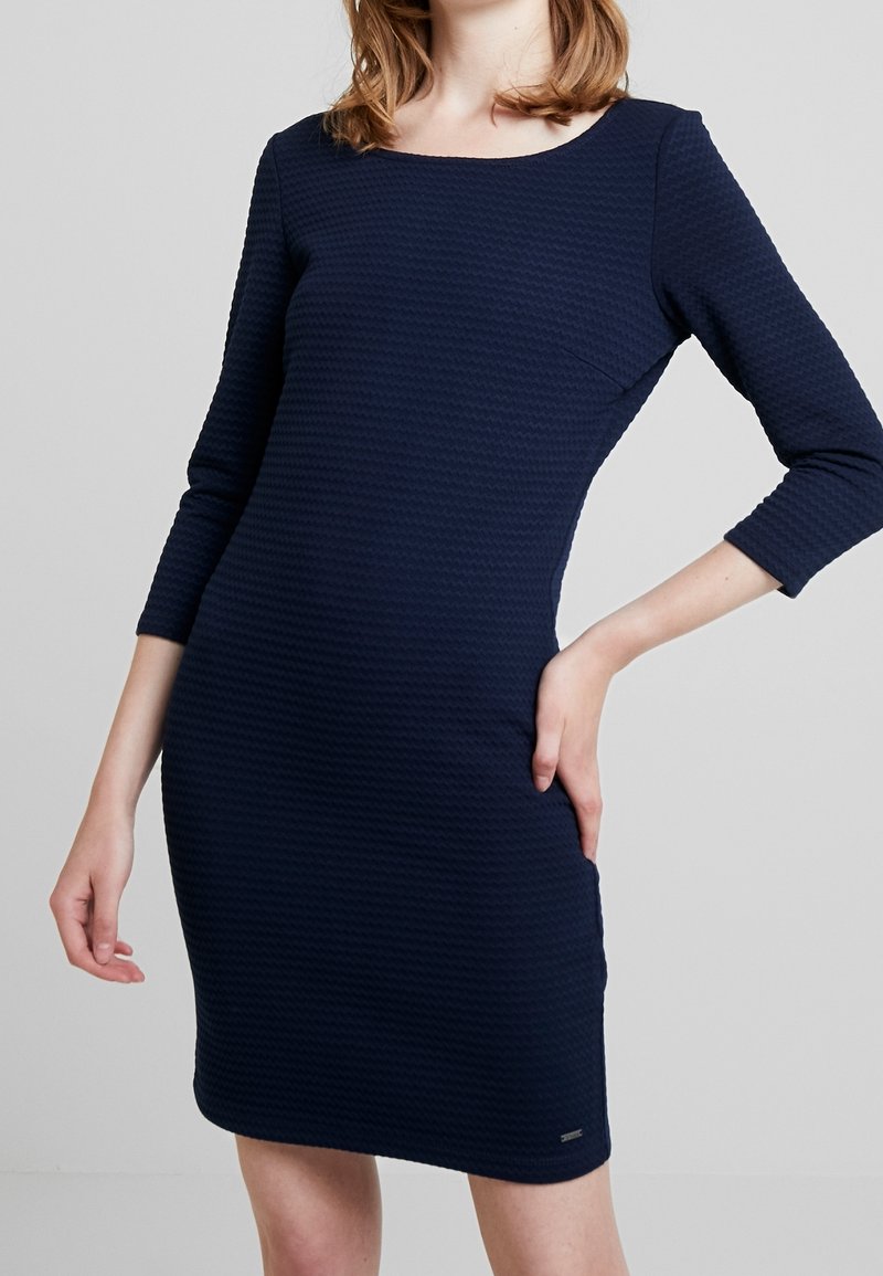 Robe bleu marine en tissu texturé, avec un col rond et des manches trois-quarts, silhouette ajustée et détail de marque subtil.