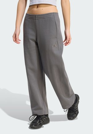 Osoba nosící volné šedé tepláky s logem Adidas, černé sportovní boty a světlý krátký top na jednobarevném pozadí.