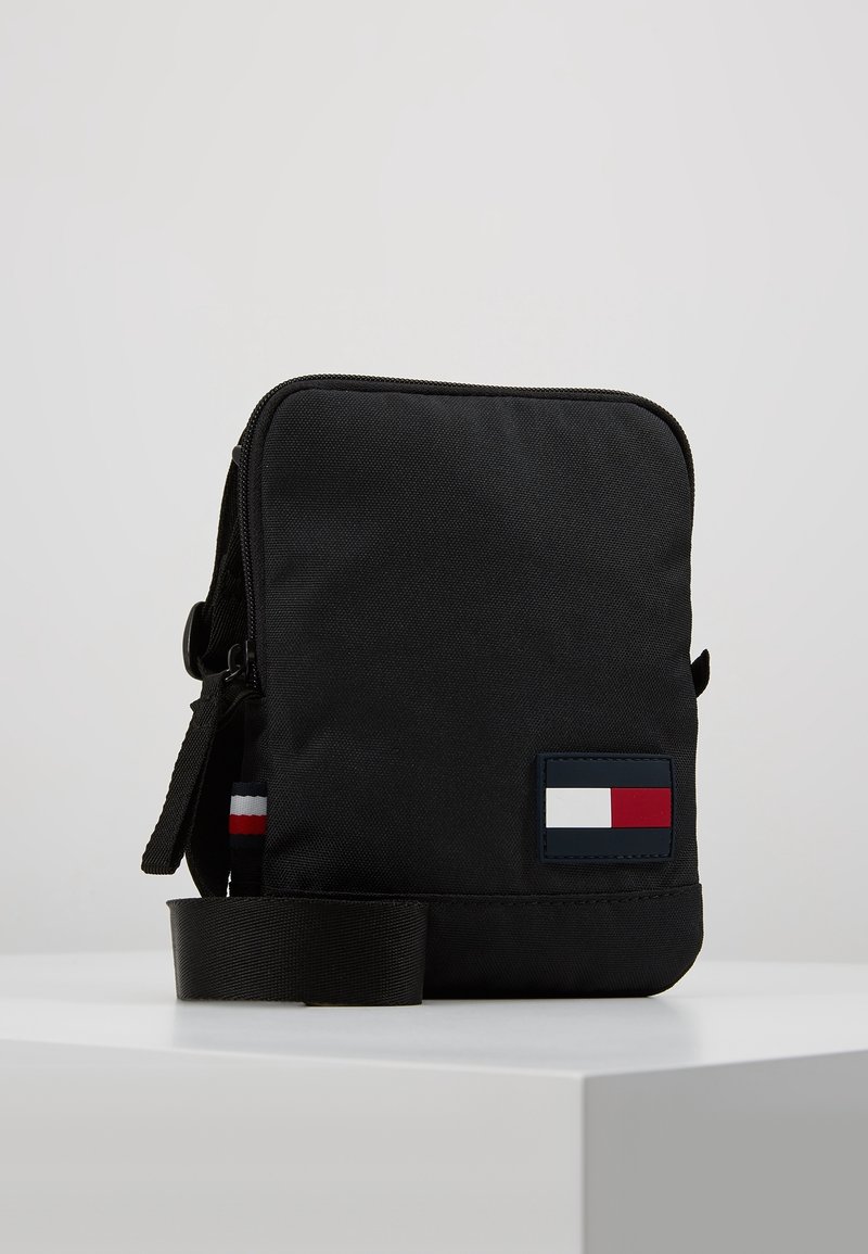 Petit sac bandoulière noir compact avec une sangle ajustable et un patch logo rectangulaire en blanc, rouge et bleu marine en bas à droite à l’avant.