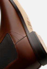 Botte en cuir marron avec un panneau latéral élastique noir, présentant une surface lisse, un bout arrondi et une semelle de couleur claire contrastante.