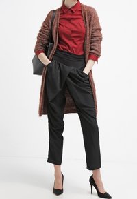 Lång brun kofta över en röd skjorta med knappar, svarta byxor med hög midja och svarta stilettklackar. Mjuka texturer, figurnära design.