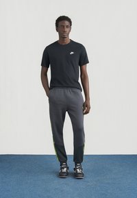 Maglietta nera Nike con piccolo logo, abbinata a pantaloni jogger grigi con strisce laterali verde neon. Indossata con scarpe sportive nere.