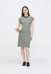 Zwart-wit gestreepte jurk tot op de knie met mouwloze cap sleeves en een ronde halslijn, gecombineerd met een lichtgekleurde handtas en zwarte loafers.