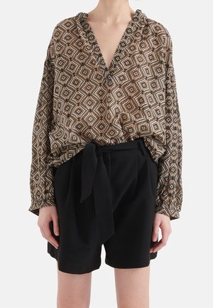 Person trägt eine braun-beige geometrisch gemusterte langärmelige Bluse, in hoch taillierte schwarze Shorts mit gebundenem Gürtel gesteckt.