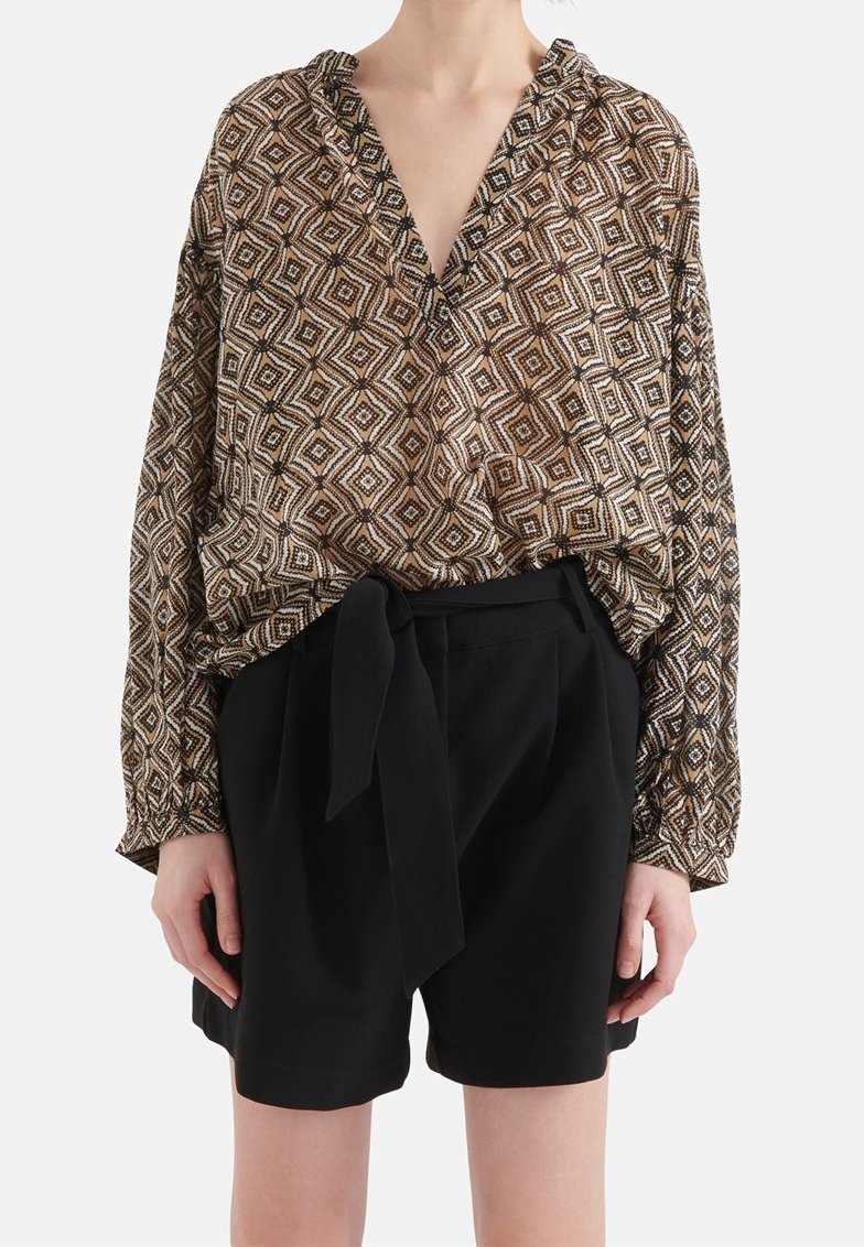 Personne portant un chemisier à manches longues à motifs géométriques marron et beige, rentré dans un short noir taille haute avec une ceinture nouée.