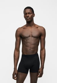 Αθλητικός άνδρας με σκουρόχρωμο δέρμα, όρθιος, φορώντας στενά μαύρα boxer briefs σε απλό φωτεινό φόντο.