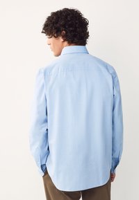 Camicia azzurro chiaro a maniche lunghe in cotone, con spalla posteriore, polsini con bottoni e orlo dritto. Tessuto liscio con dettagli di design minimi.