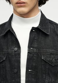 Homme portant un col roulé blanc sous une veste en jean noire avec des fermetures à boutons et des poches poitrine, coupée au niveau des lèvres.