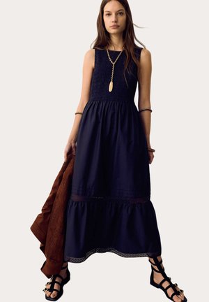 Frau trägt ein ärmelloses, navy Maxi-Kleid, goldene Halskette, Armbänder, schwarze Riemchensandalen und hält eine braune, strukturierte Jacke in der linken Hand.