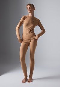 Woman standing barefoot wearing fitted tan long-sleeve top and leggings against a plain gray background, posing with hands on hips.