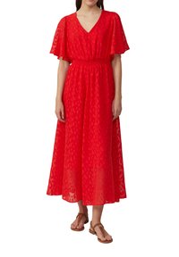 Vrouw draagt een rode midi-jurk met flapmouwen en een aansluitende taille, gecombineerd met bruine platte sandalen, staand tegen een witte achtergrond.