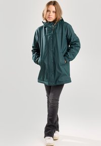 Teal waterproof jacket with a hood, button closure, elastic cuffs, and side pockets, paired with dark jeans and white sneakers.
