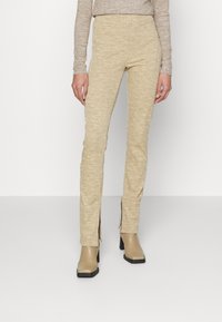 Béžové kalhoty s texturou a bočními rozparky, s přiléhavým střihem a elastickým pasem, doplněné béžovými botami na podpatku.