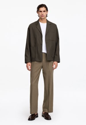 Uomo con giacca verde oliva scuro abbottonata, t-shirt bianca, pantaloni dritti marrone chiaro e mocassini marrone scuro che sta contro uno sfondo bianco.