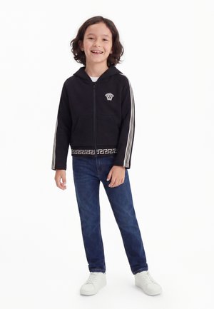 Kind mit schulterlangem Haar, das einen schwarzen Hoodie mit Reißverschluss und weißen Streifen, blaue Jeans und weiße Sneakers trägt und dabei lächelt.