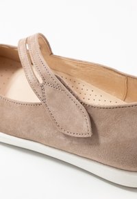 Zapato de ante beige con punta redondeada, que presenta detalles de costura y una correa con lazo. El interior tiene perforaciones para una mejor transpirabilidad.