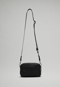 Černá crossbody taška vyrobená z měkkého materiálu, obdélníkového tvaru, se zipovým uzávěrem, nastavitelným popruhem a stříbrnými kovovými detaily.