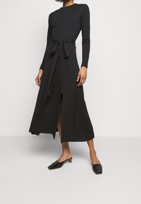 Abito nero a maniche lunghe con vita legata, dotato di uno spacco frontale e una silhouette fluida. Abbinato a mocassini neri.