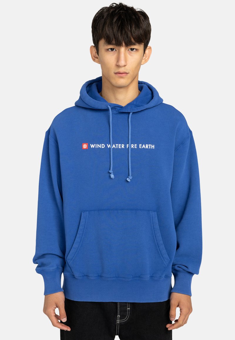 Giovane vestito con una felpa blu con tasca frontale e il testo "VENTO ACQUA FUOCO TERRA" sul petto, in piedi davanti a uno sfondo neutro.