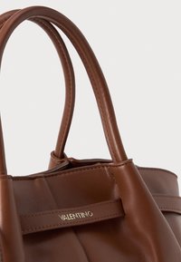 Mala de mão em pele castanha com duas alças, apresentando um detalhe em logótipos dourados e costuras subtis. Textura suave, design estruturado.