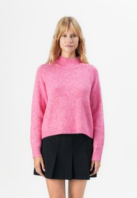 Mulher vestindo um suéter felpudo rosa-choque e uma saia mini plissada preta, de pé contra um fundo branco.