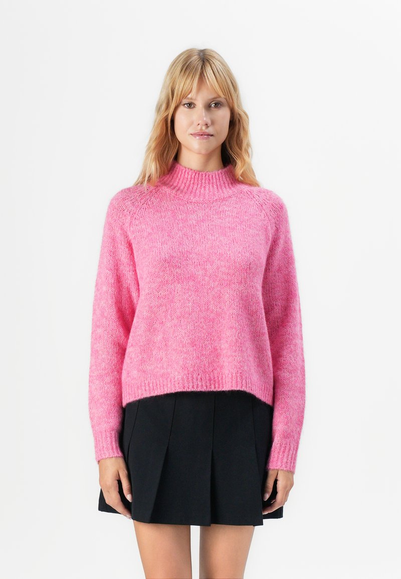 Mulher vestindo um suéter felpudo rosa-choque e uma saia mini plissada preta, de pé contra um fundo branco.