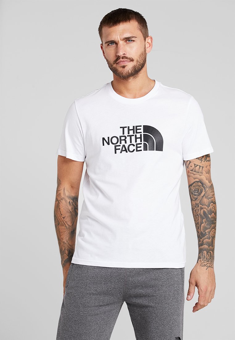 Valge puuvillane T-särk, millel on silmapaistev must logo, mis näitab tekstiga "THE NORTH FACE". Mudel kannab halli pükse ja tema kätes on nähtavad tätoveeringud.
