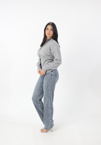 Cardigan grigio con scollo a V, design aderente, indossato sopra una maglia scura, abbinato a jeans dritti blu chiaro e sandali con tacco trasparente.