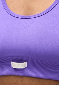 Primer plano de una persona con un sujetador deportivo morado con una etiqueta blanca que dice "BORG" en la banda acanalada debajo del pecho.