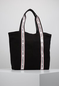 Svart tote-väska med breda vita tyghandtag som pryder upprepande "KARL LAGERFELD"-text i svart, placerad på en vit piedestal.