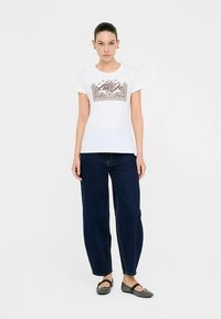 White t-shirt with decorative "Liu Jo" text and graphic design, paired with dark blue wide-leg jeans and gray shoes.