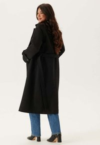 Cappotto di lana nera con vita legata, maniche lunghe e tasche laterali. Abbinato a jeans blu a gamba dritta e scarpe con tacco nere.