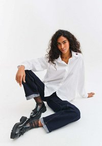 Žena s kudrnatými vlasy sedí na podlaze, má na sobě bílou košili na knoflíky, tmavé ohrnuté džíny, průsvitné třpytivé ponožky a černé mokasíny.