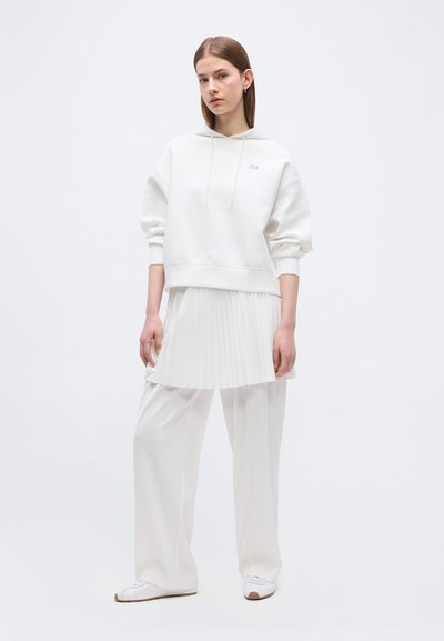 Jeune femme portant un sweat à capuche blanc, une jupe plissée superposée à un pantalon large, et des baskets blanches, se tenant devant un fond uni.