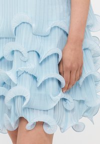 Light blue dress featuring layered, ruffled hem with textured pleats and a smooth finish. Hand visible holding fabric near the waist.