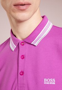 Polo à manches courtes en tissu rose vif, avec un col rayé blanc, trois boutons et un logo blanc sur la poitrine gauche.