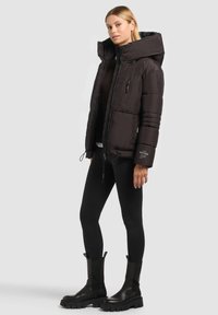 Doudoune noire avec col montant et fermeture éclair, présentant des coutures verticales, des poches zippées et un ourlet à cordon ajustable. Assortie à des leggings noirs et des bottes noires épaisses.