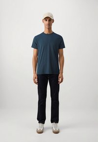 Bavlnené tričko tyrkysovej farby v kombinácii s tmavými džínsami a bielymi teniskami s béžovými akcentmi. Model má na hlave svetlobéžovú čiapku. Jednoduchý dizajn, ležérny strih.