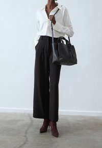 Žena v bílé blůze, černých kalhotách s volnými nohavicemi, hnědých botách na podpatku a s velkou černou koženou kabelkou přes rameno.