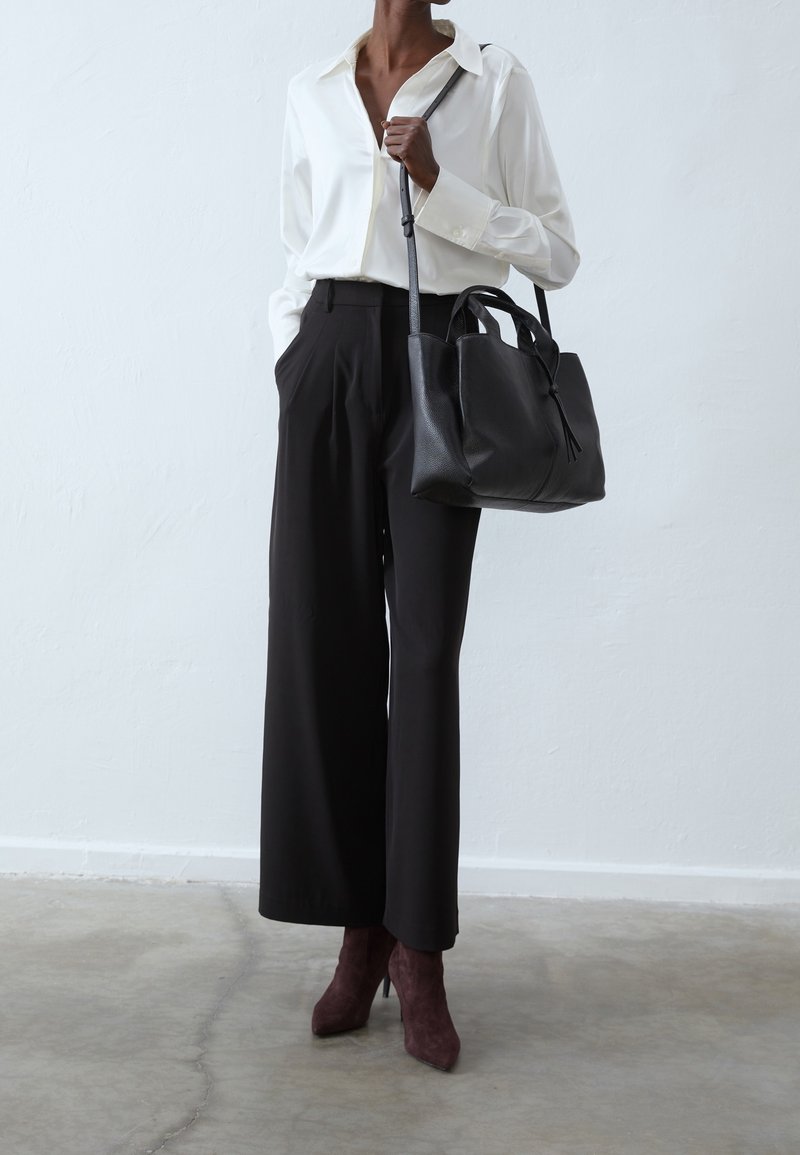 Žena v bílé blůze, černých kalhotách s volnými nohavicemi, hnědých botách na podpatku a s velkou černou koženou kabelkou přes rameno.