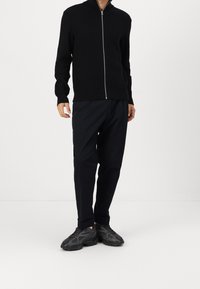 Pull zippé noir à côtes avec un col haut, associé à un pantalon sombre et des baskets noires imposantes, offrant une coupe moderne et décontractée.