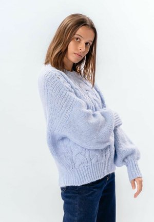 Jonge vrouw draagt een lichtblauwe oversized kabelgebreide trui en donkere jeans, poserend tegen een effen witte achtergrond.